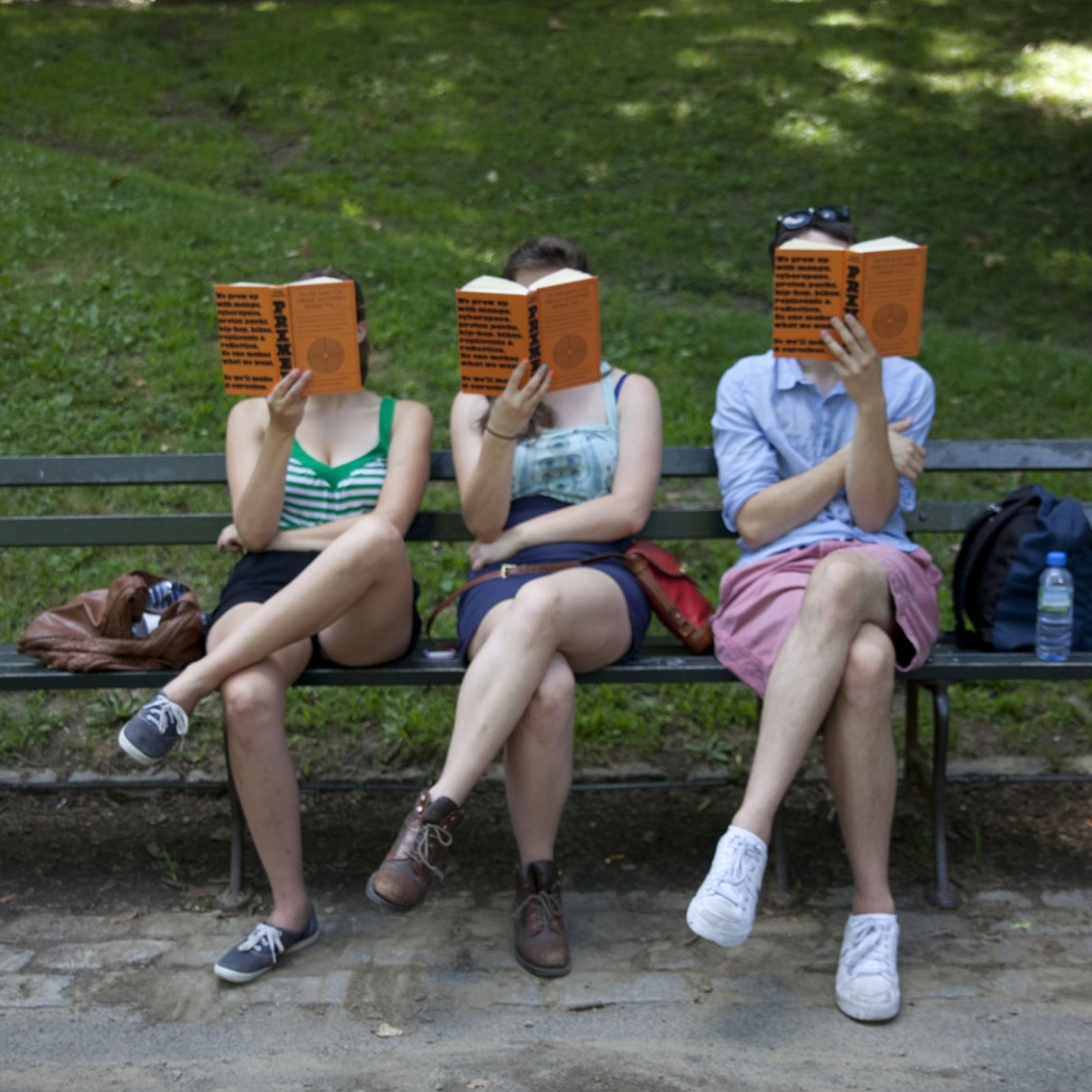 Three students sitting on a park bench, each reading the same orange-covered book.