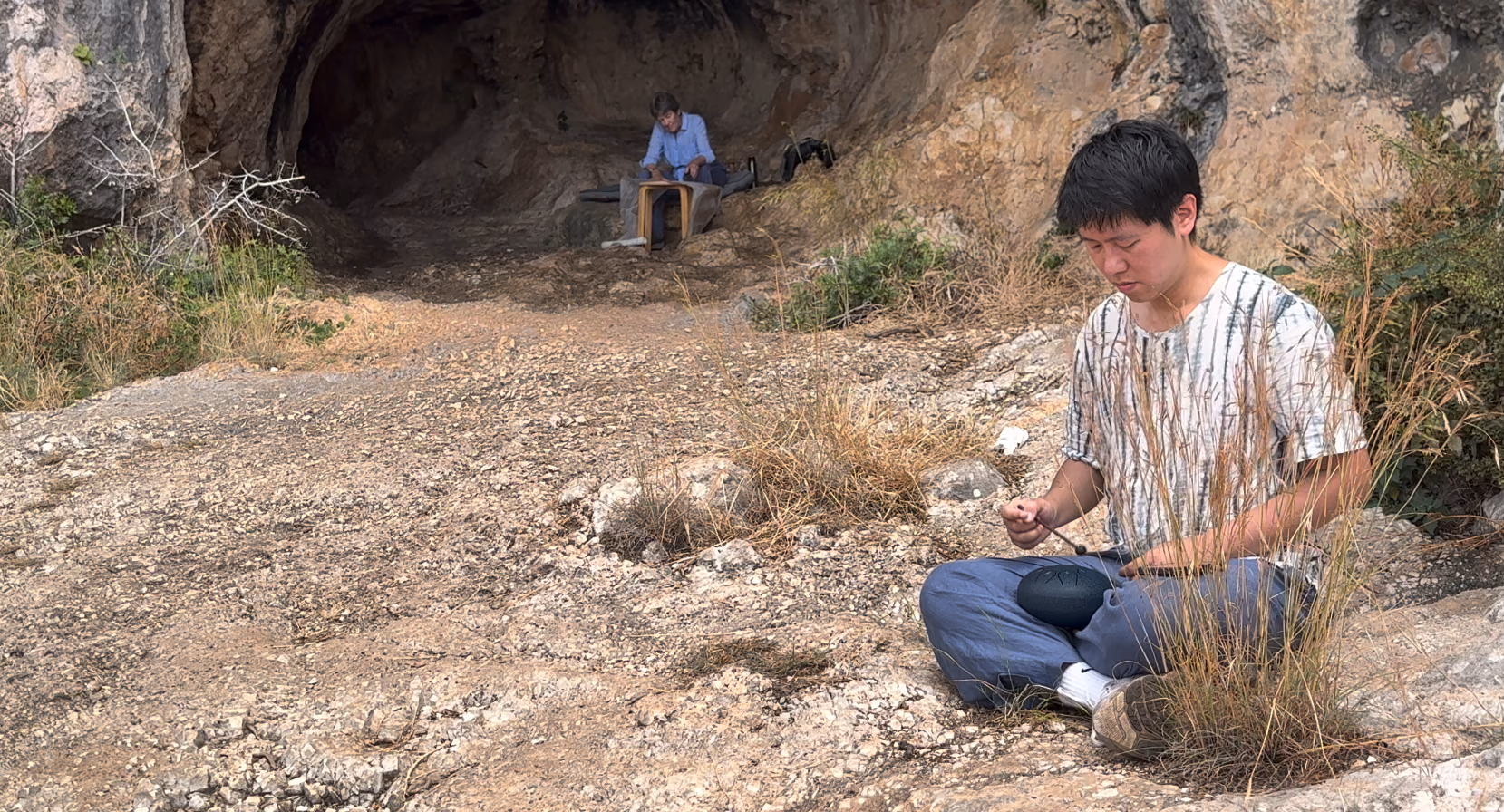 A limestone cave mouth; one performer sits at its entrance and another waits in the scrub outside.