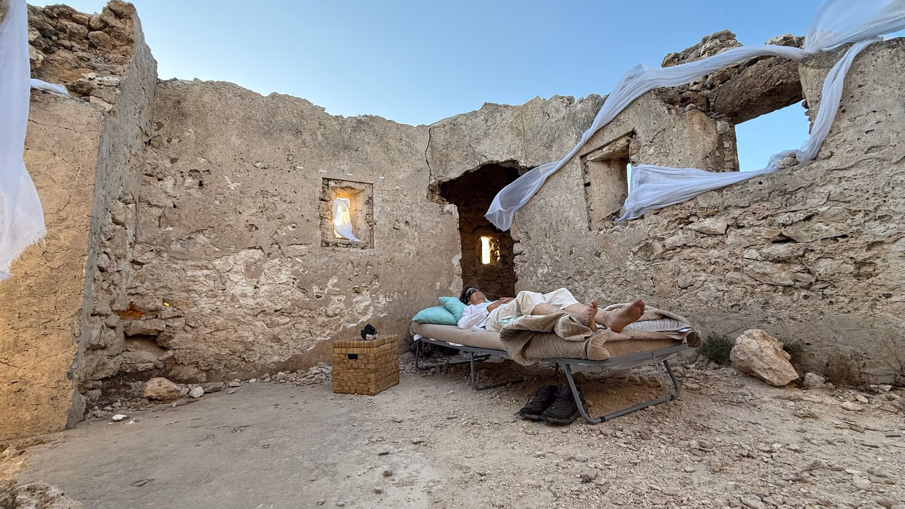 A made bed stands in the roofless ruin of a stone building, white cloth lifting from the walls at dusk.