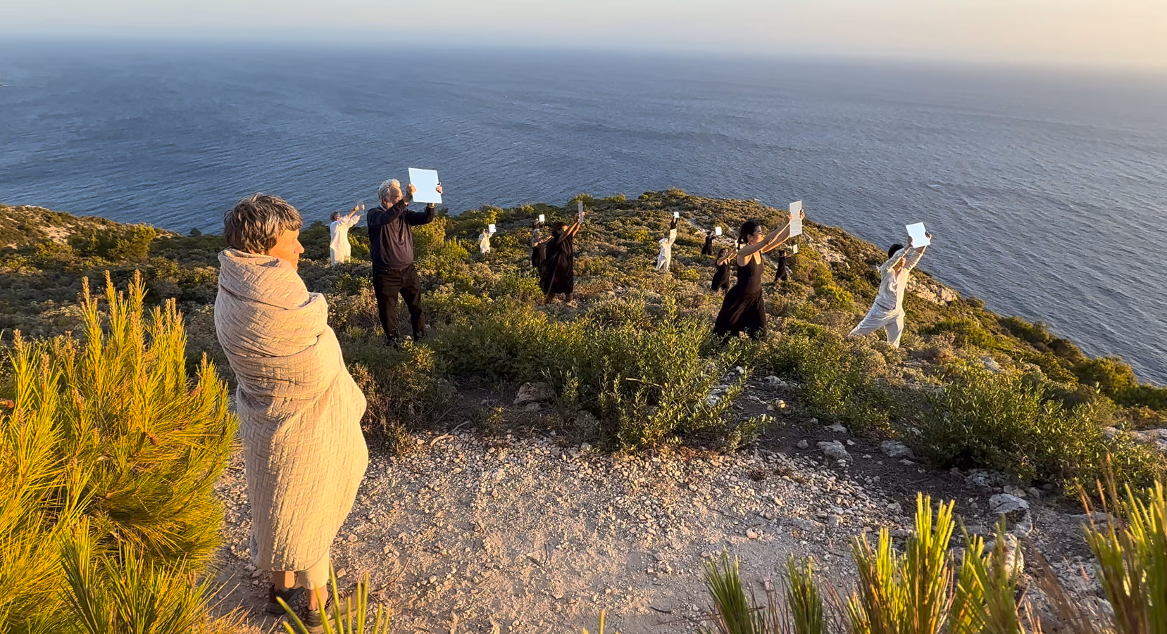 On a clifftop above the sea at sunset, Roz stands wrapped in a blanket while dancers hold large mirrors up to the light.
